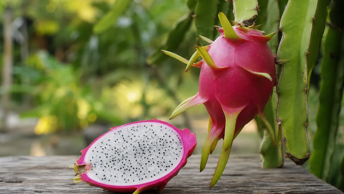 A ripe magenta dragon fruit with green-tipped scales beside a cut half showing white flesh with black seeds on a wooden table